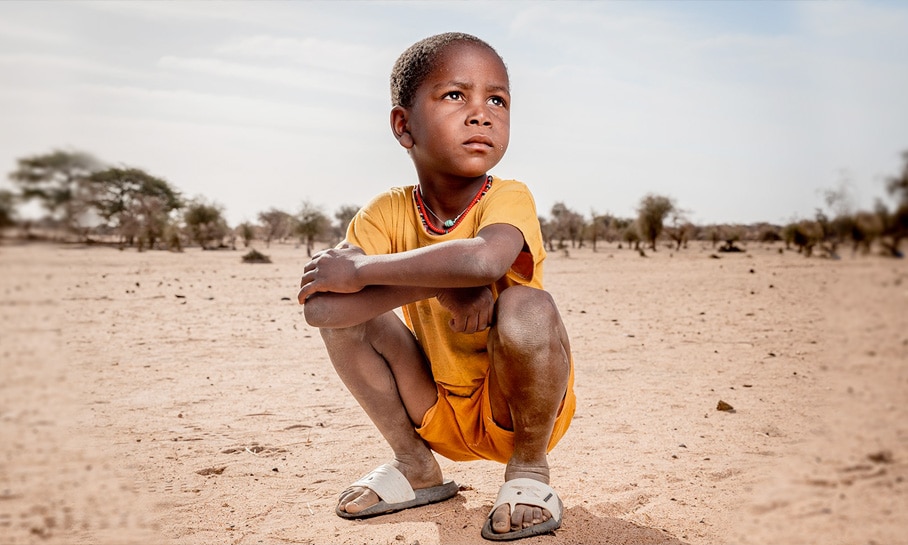 Enfant accroupi dans un paysage désertique, portant un t-shirt jaune et un collier traditionnel, symbole de résilience face aux défis climatiques en Afrique.