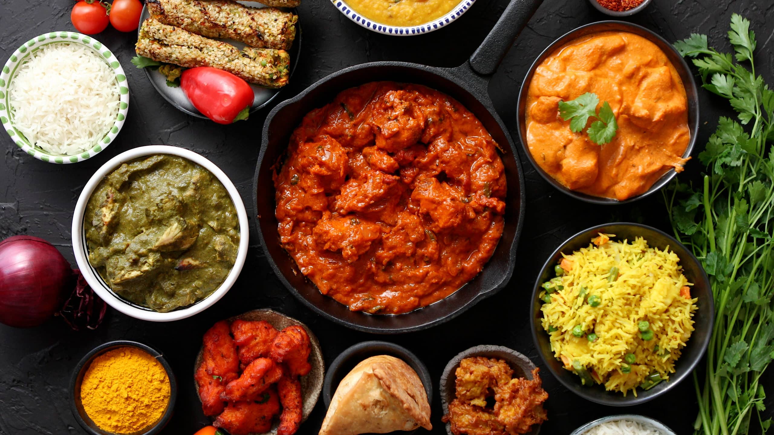 Un assortiment de plats indiens traditionnels incluant du riz basmati, des currys, des brochettes végétariennes, des tomates, des épices, et des légumes frais, disposés sur une table en marbre noir.