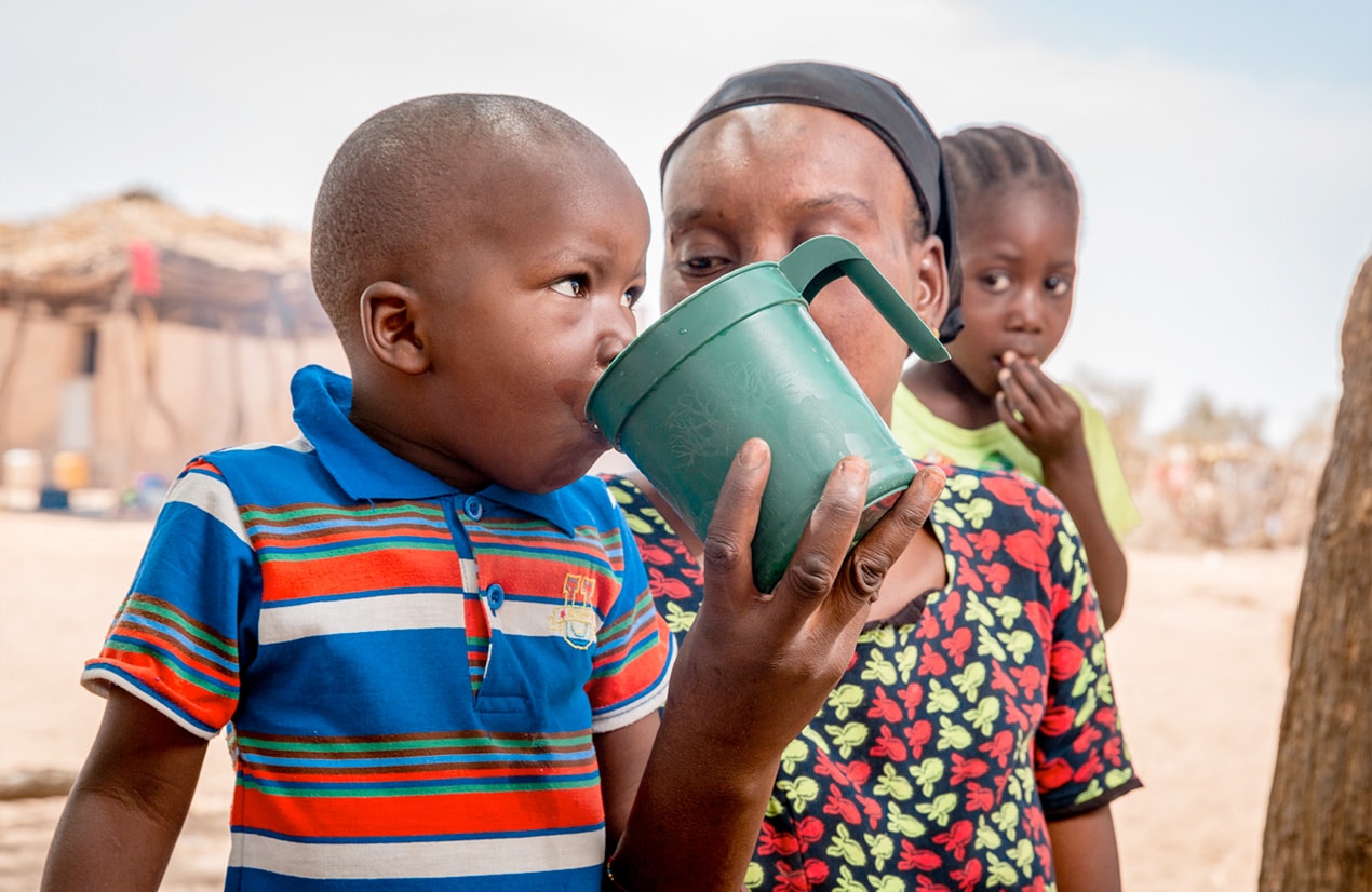 Un jeune garçon sénégalais en chemise rayée de couleurs vives boit dans une grande tasse verte tenue par une femme, avec une petite fille en arrière-plan.