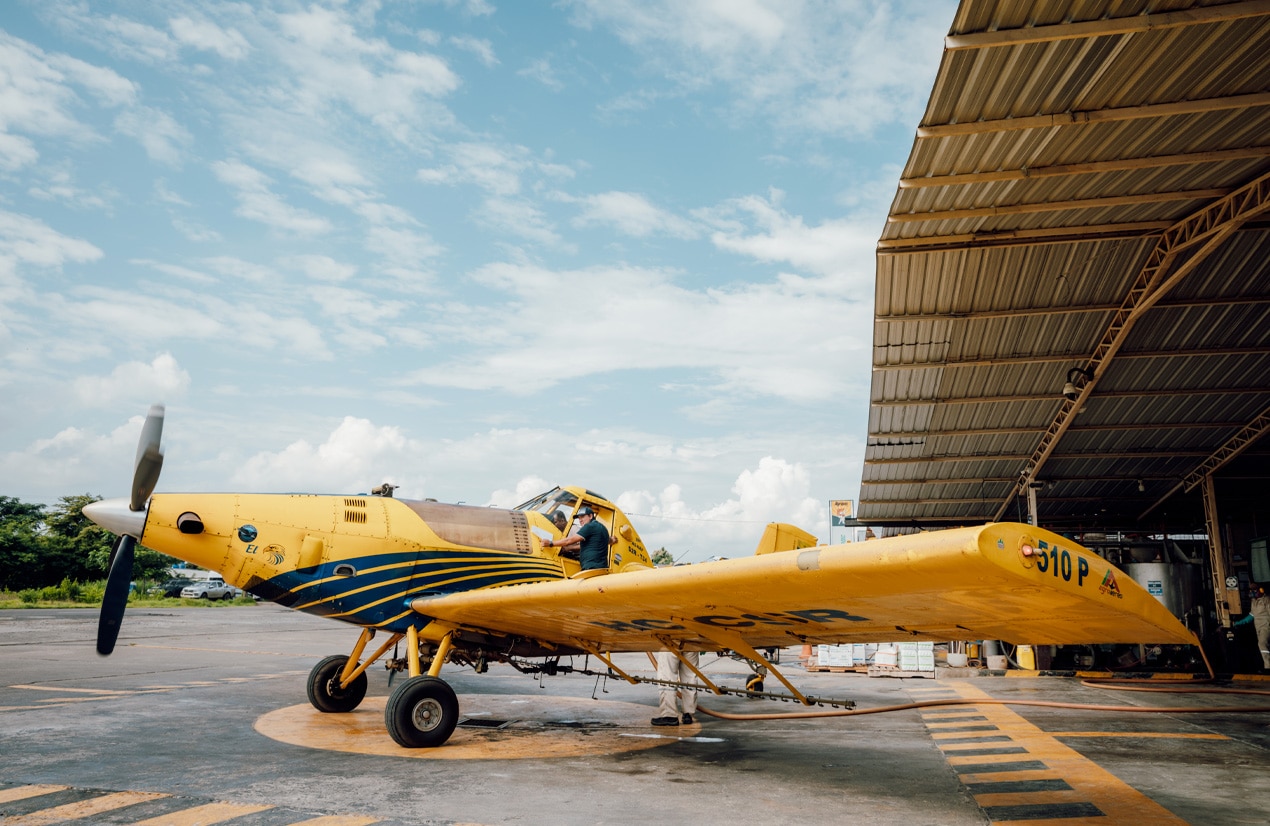 Un avion jaune à hélice, de type agricole ou de pulvérisation, est stationné sur une piste d'aviation. Un homme en chemise bleue est debout à côté de l'avion, occupé à faire des vérifications ou des préparations avant le vol. L'avion est partiellement couvert par un hangar métallique, et le ciel est dégagé avec quelques nuages.