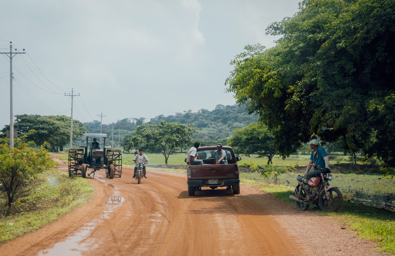 Scène rurale avec une route en terre traversant une campagne verdoyante. Un tracteur, une moto et une camionnette avec plusieurs personnes à bord sont visibles sur la route. En arrière-plan, des collines couvertes de végétation dense. Un homme assis sur une moto est stationné à l'ombre d'un grand arbre à droite de l'image.