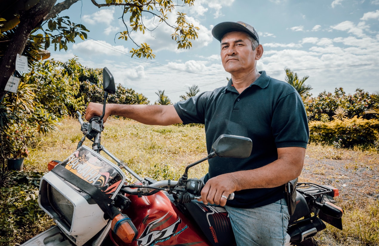 Un homme en chemise polo noire et casquette repose sur une moto rouge de marque Yamaha. En arrière-plan, un paysage rural verdoyant avec des arbres et un ciel partiellement nuageux. L'homme regarde droit devant avec une expression sérieuse.