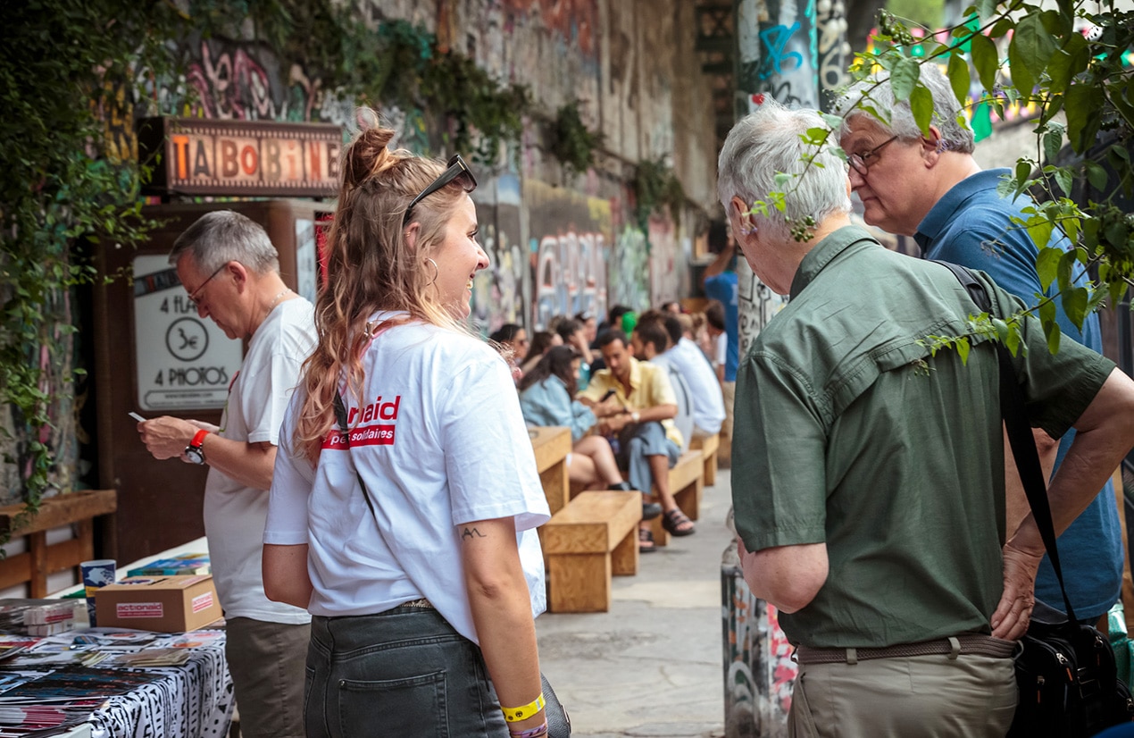 Des personnes discutent et interagissent lors d'un événement en plein air. Une femme portant un t-shirt blanc avec le logo 'ActionAid' parle à deux hommes plus âgés. En arrière-plan, on peut voir d'autres participants assis sur des bancs en bois, le long d'un mur recouvert de graffitis. La scène se déroule dans un espace animé et convivial, avec de la végétation visible au premier plan.
