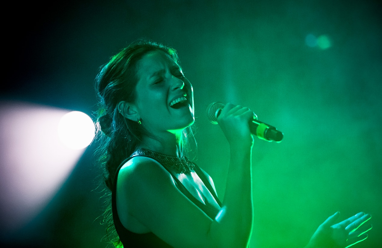 Une femme chante avec passion dans un micro sur scène, éclairée par une lumière verte intense. Ses cheveux sont attachés en arrière et elle porte des boucles d'oreilles. L'arrière-plan est sombre avec un éclairage dramatique.