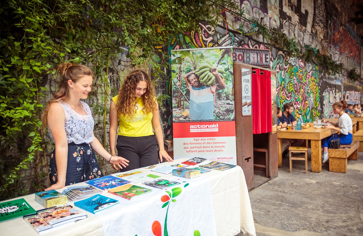 Deux jeunes femmes se tiennent derrière une table d'exposition recouverte de brochures et de documents. Elles sont dans un espace extérieur décoré de graffitis et de verdure. Un poster d'ActionAid est visible en arrière-plan, mettant en avant la mission de l'organisation pour les droits humains à travers le monde. En arrière-plan, des gens sont assis à des tables en train de manger et discuter.