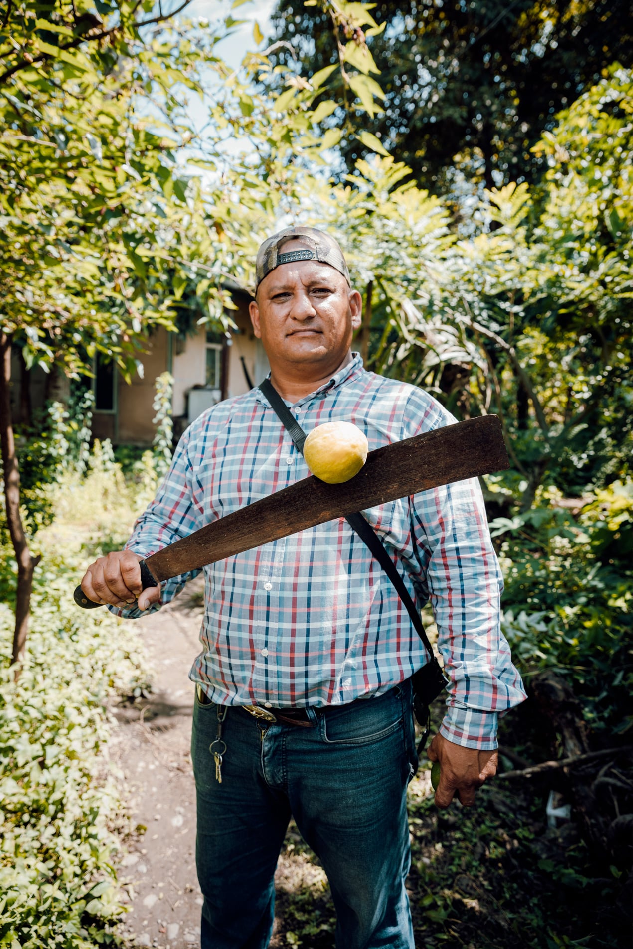Portrait de Presley portant une chemise à carreaux et casquette à l'envers, tenant une grande machette avec un fruit jaune posé dessus, dans un jardin verdoyant.