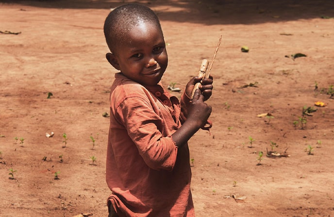 Jeune garçon centrafricain souriant, tenant un jouet fait maison en plein air sur un fond de terre, illustrant l'innocence de l'enfance et la créativité dans un contexte rural africain.