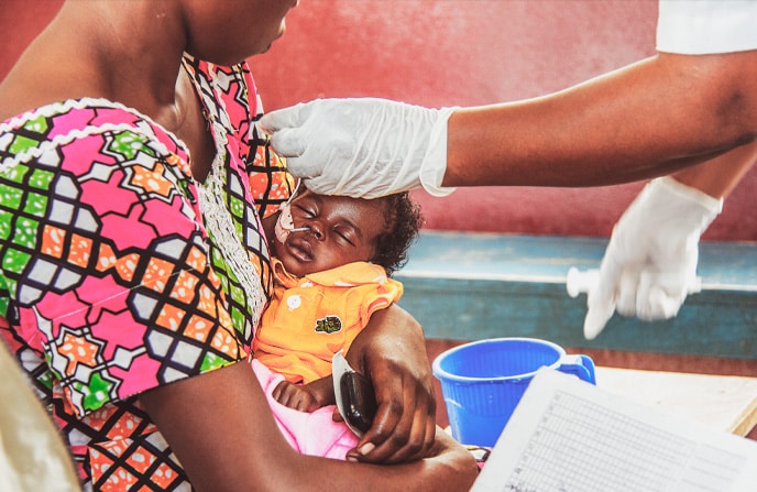 Mère centrafricaine tenant son bébé lors d'un examen médical par une infirmière portant des gants, dans un centre de santé avec des outils médicaux et des documents visibles, illustrant les soins de santé en contexte communautaire.