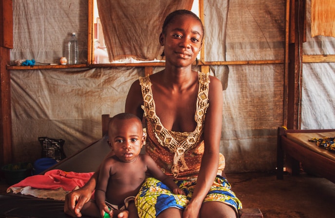 Mère centrafricaine souriante assise avec son jeune enfant, assis sur un lit sommaire dans l'intérieur rustique de l'hôpital pédiatrique de Bangui, exprimant la complicité et l'amour familial, avec un arrière-plan de tentures et d'objets du quotidien.