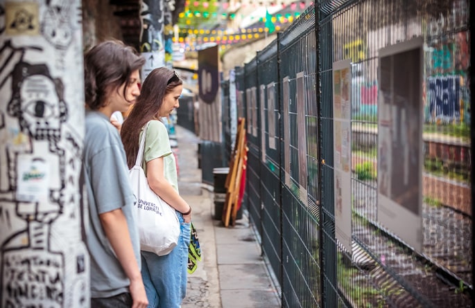 Deux jeunes femmes observent attentivement une exposition extérieure de photographies et d'affiches informatives accrochées sur un grillage. Elles semblent plongées dans la contemplation des œuvres. La scène se déroule dans une allée ornée de graffitis colorés et de guirlandes lumineuses, ce qui ajoute une atmosphère artistique urbaine à l'expérience de l'exposition.