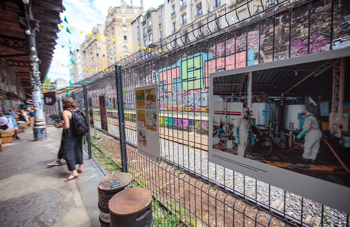 Une allée urbaine accueille une exposition photo en plein air, avec des images de grand format fixées sur un grillage. Les photos montrent des scènes variées, dont certaines semblent liées à la nutrition et à l'agriculture. Une femme se tient à distance, examinant les œuvres. L'allée est bordée de murs couverts de graffitis colorés et ornée de guirlandes de drapeaux, créant un environnement dynamique et artistique pour l'exposition.