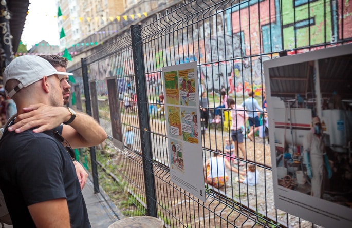 Deux hommes regardent une série de panneaux d'exposition affichés sur un grillage dans une ruelle. Les panneaux présentent des informations visuelles, photographies et illustrations. À l'arrière-plan, l'allée est animée par des personnes et des guirlandes de fanions multicolores, donnant à la scène une ambiance festive et communautaire.