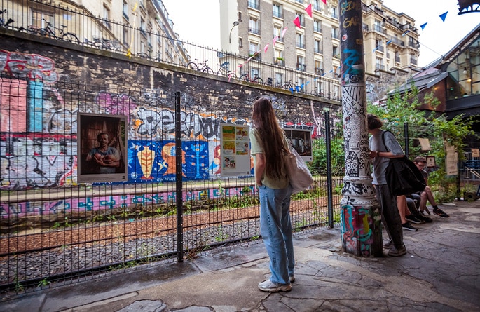 Une ruelle vivante avec des spectateurs observant des photographes et illustrations exposées sur un grillage. Une femme au premier plan regarde attentivement une image encadrée, tandis qu'une autre personne s'appuie sur un poteau couvert de graffitis colorés. Des fanions suspendus ajoutent une touche festive à la scène urbaine, pleine de tags et d'expressions artistiques sur les murs en arrière-plan.