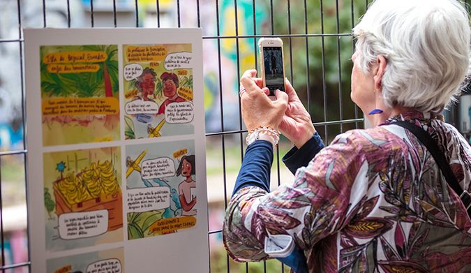 Une femme aux cheveux gris photographie un panneau d'exposition à l'aide de son smartphone. Le panneau, affiché sur une grille métallique, présente une bande dessinée de Blanche Sabbah, avec plusieurs cases illustrant des discussions sur la nutrition. La femme est vue de dos, concentrée sur la capture de l'image.